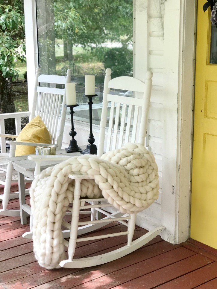 yellow door with 3 white rocking chairs on porch with hanging bats and cobwebs wiht pumpkins and whte chunky blanket on one rocking chair